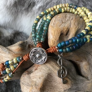 May include: A multi-strand bracelet with blue, green, and yellow beads. The bracelet has a silver charm in the shape of a treble clef and a silver button with a floral design. The bracelet is tied with brown leather cord.