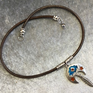 May include: A dark brown leather cord necklace featuring a silver-toned crescent moon pendant. The pendant is decorated with blue and orange enamel accents and a small dangling silver feather charm. The necklace includes silver metal end caps.