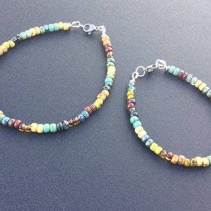 May include: Two colorful beaded bracelets with silver clasps. The bracelets are made with small, round beads in shades of blue, green, yellow, and brown.