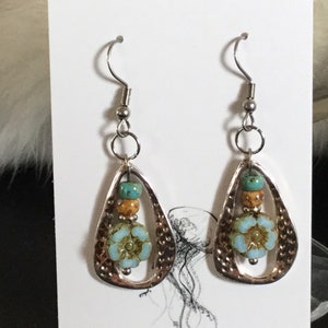 May include: A pair of silver dangle earrings with a teardrop-shaped design. The earrings feature a blue and green flower charm with brown and gold beads.