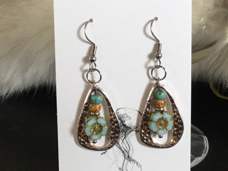 May include: A pair of silver dangle earrings with a teardrop-shaped design. The earrings feature a blue and green flower charm with brown and gold beads.