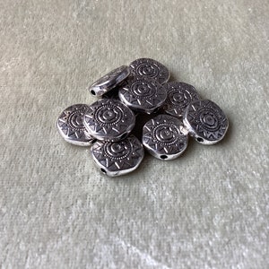 May include: A pile of silver metal beads with a circular design. The beads have a hole in the center for stringing.