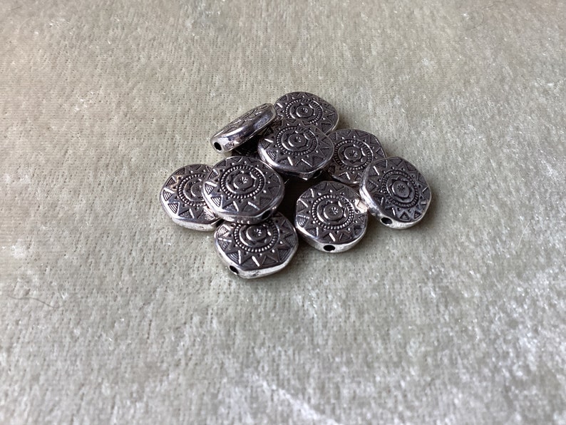 May include: A pile of silver metal beads with a circular design. The beads have a hole in the center for stringing.