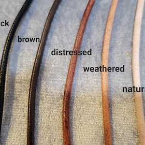 K&ouml;nnte beinhalten: Nahaufnahme von f&uuml;nf Lederkordeln in verschiedenen Farben: Schwarz, Braun, Distressed, Weathered und Natur. Jede Kordel ist mit ihrem jeweiligen Farbnamen beschriftet. Die Kordeln sind nebeneinander angeordnet und zeigen die verschiedenen Schattierungen und Texturen.