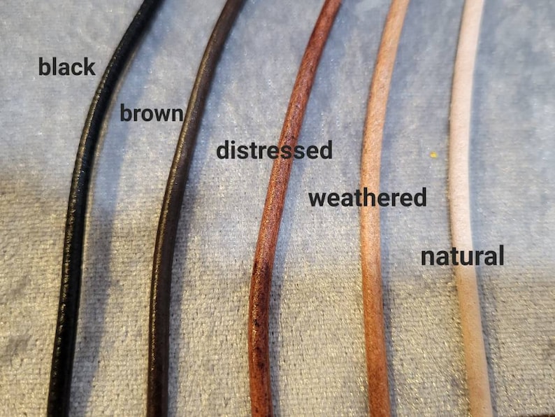 K&ouml;nnte beinhalten: Nahaufnahme von f&uuml;nf Lederkordeln in verschiedenen Farben: Schwarz, Braun, Distressed, Weathered und Natur. Jede Kordel ist mit ihrem jeweiligen Farbnamen beschriftet. Die Kordeln sind nebeneinander angeordnet und zeigen die verschiedenen Schattierungen und Texturen.