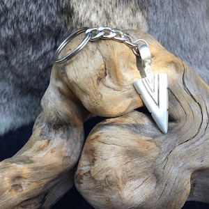 May include: Silver arrowhead keychain with a silver chain and a silver ring. The keychain is resting on a piece of driftwood.