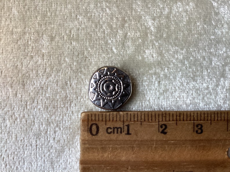 May include: A silver-toned button with a decorative design featuring a sun and moon motif. The button is approximately 1 cm in diameter.