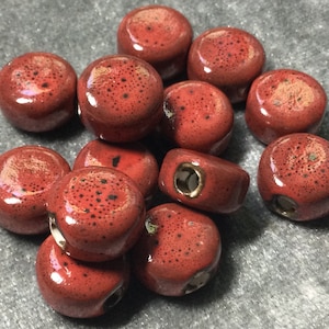 May include: A group of 12 red ceramic beads with black speckles. The beads have a hole through the center for stringing.