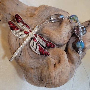 May include: A red and silver dragonfly charm with crystal accents hangs from a beaded chain with iridescent glass beads.