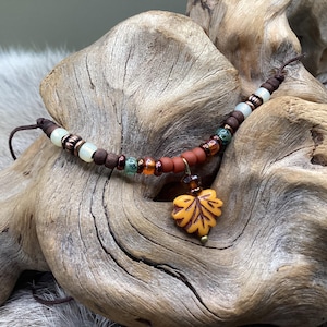 May include: A handcrafted necklace featuring a leaf pendant. The necklace has a brown cord with various beads in shades of brown, green, and orange. The leaf pendant is yellow and orange. The necklace is displayed on a piece of driftwood.