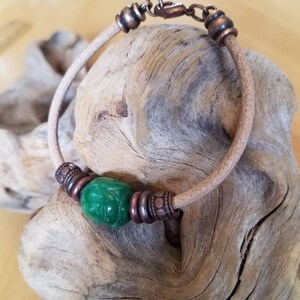 May include: A leather bracelet with a green jade bead and copper accents. The bracelet is on a piece of driftwood.