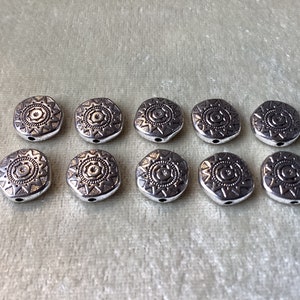 May include: Ten silver-toned metal beads with a geometric design. Each bead features a circular pattern with a star in the center.