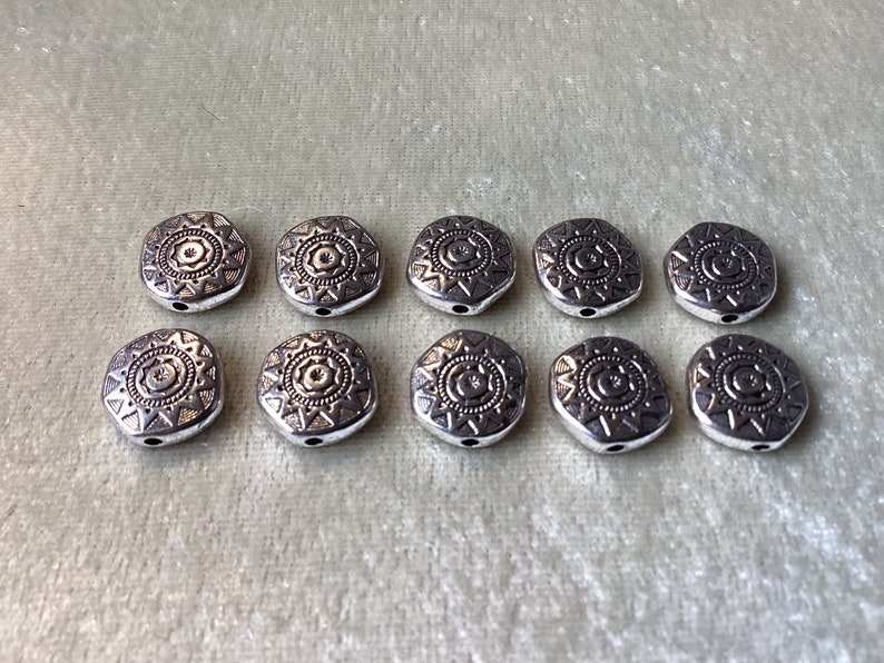May include: Ten silver-toned metal beads with a geometric design. Each bead features a circular pattern with a star in the center.