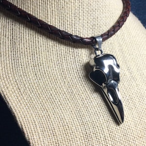 May include: A silver crow skull pendant on a brown leather cord necklace.