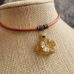 May include: A brown leather choker necklace with a gold flower pendant. The pendant has a textured surface and a raised center.