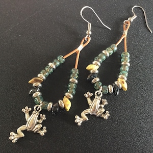 May include: A pair of dangle earrings with a frog charm. The earrings are made with green, black, and gold beads and have a silver wire.