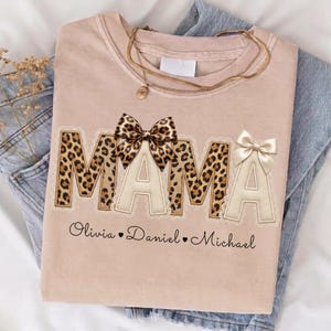 Pode incluir: Uma t-shirt bege claro com a palavra MAMA em letras grandes com padrão de leopardo e um laço cor de creme. Abaixo do texto, os nomes Olivia, Daniel e Michael estão impressos em letra cursiva preta, separados por pequenos corações.