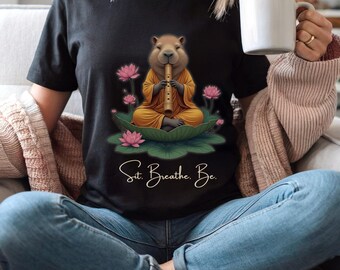 May include: Black t-shirt featuring a capybara playing a flute while seated on a lily pad, surrounded by pink lotus flowers. The capybara wears a yellow robe. The text "Sit. Breathe. Be." is written in cursive below the image.