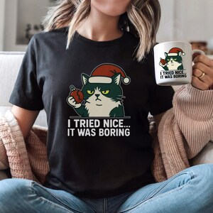 Może przedstawiać: Czarny t-shirt z wrednym kotem w czapce Świętego Mikołaja trzymającym bombkę. Pod kotem wydrukowano napis "I TRIED NICE... IT WAS BORING". Widoczny jest również pasujący kubek z tym samym wzorem.