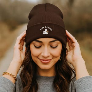 Pode incluir: Um gorro castanho escuro com o texto bordado "BOOK DRAGON" e um gráfico branco de um dragão segurando um livro. O gorro é feito de um material de malha canelada.