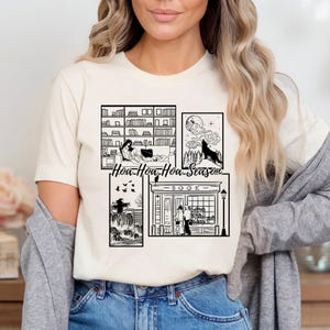 May include: Cream-colored t-shirt featuring a black and white graphic design with the text "Hoa-Hoa-Hoa-Season". The design includes illustrations of a bookshelf, a wolf howling at the moon, a witch, and a bookstore.