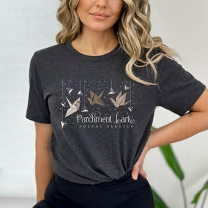 May include: Dark gray t-shirt featuring a design with three origami cranes in white and brown tones, suspended from strings with geometric shapes. The text "Parchment Lark Postal Service" is below the cranes.
