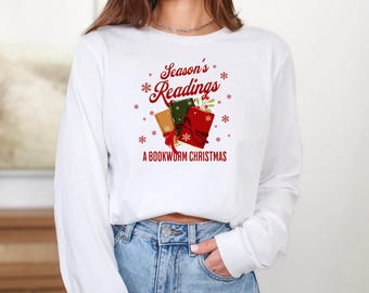 Può includere: Maglietta bianca a maniche lunghe con grafica natalizia. Il design presenta tre libri impilati con fiocchi rossi, il testo "Season's Readings" e "A Bookworm Christmas" in rosso e verde, con fiocchi di neve.