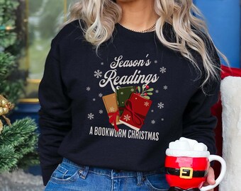 Può includere: Una felpa blu navy con la scritta "Season's Readings" e "A Bookworm Christmas" stampata in bianco. La grafica presenta tre libri con decorazioni festive. Viene tenuta una tazza a tema Babbo Natale con un manico bianco.