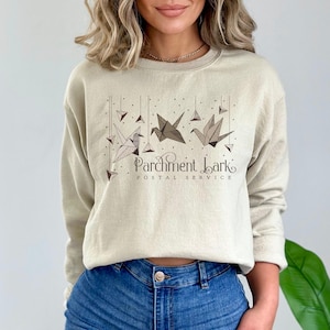 Könnte beinhalten: Beiges Sweatshirt mit einer Grafik, die Origami-Kraniche und geometrische Formen zeigt. Der Text "Parchment Lark Postal Service" ist darunter gedruckt. Das langärmlige Oberteil ist aus weichem Material.