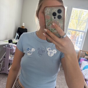 May include: Pale blue crop top with short sleeves. The shirt has three Mickey Mouse head appliqués in floral, gingham, and bow patterns. The model is wearing a striped skirt.