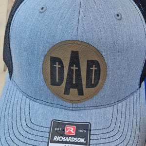 May include: A gray and black trucker hat with a brown leather patch that says "DAD" in black letters with a gold cross on each side of the word. The hat has a black mesh back and a Richardson logo with the text "EST. 1970 ORIGINAL 112 TRUCKER ADJUSTABLE".