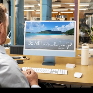 May include: A light blue desktop computer monitor displaying a beach landscape with the white text Be Still and know that I Am God Psalm 46:10. The monitor sits on a wooden desk with a white keyboard, a white mouse, and a white water bottle.