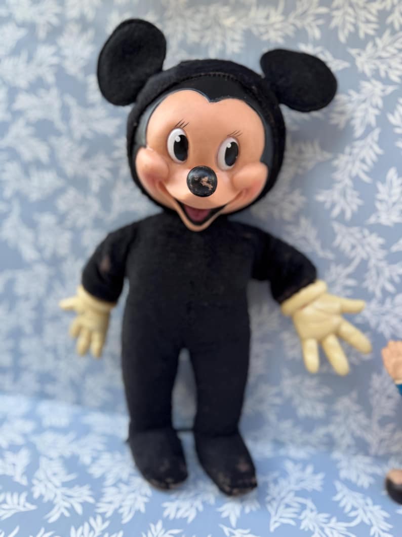 May include: Vintage Mickey Mouse doll in a black jumpsuit with a smiling face, large black ears, and cream-colored gloves. The doll's face is tan with black eyes and a black nose. A classic Disney collectible.
