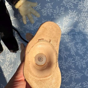 May include: A vintage doll's head, featuring a light tan complexion and a circular opening with a mesh insert. The doll's hand, with a white glove, is visible in the background. The doll's head is held against a blue background with a white leaf pattern.