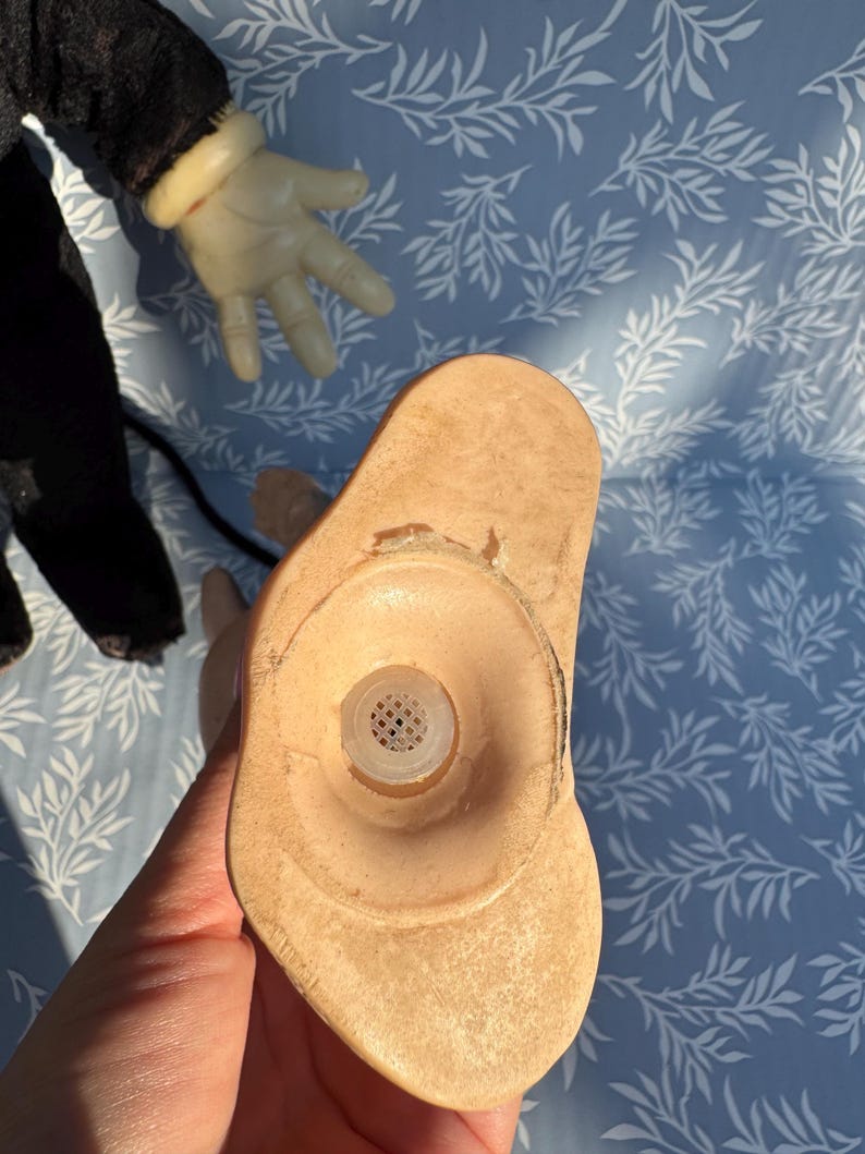 May include: A vintage doll's head, featuring a light tan complexion and a circular opening with a mesh insert. The doll's hand, with a white glove, is visible in the background. The doll's head is held against a blue background with a white leaf pattern.