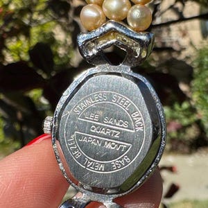 May include: A vintage Lee Sands quartz watch with a stainless steel back and metal bezel. The watch features a silver-toned case and a strand of light-colored pearls. The watch is marked "Japan Movt".