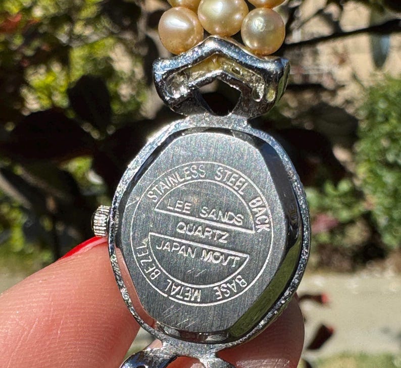May include: A vintage Lee Sands quartz watch with a stainless steel back and metal bezel. The watch features a silver-toned case and a strand of light-colored pearls. The watch is marked "Japan Movt".