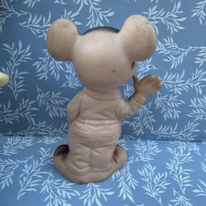 May include: Vintage, light pink mouse figurine with large ears, wearing a light pink outfit with a black head. The figurine is posed with one arm raised. The background is a blue surface with a white leaf pattern.