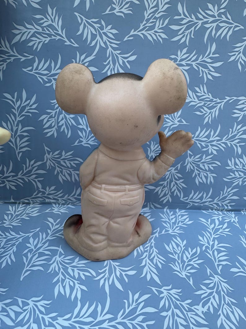 May include: Vintage, light pink mouse figurine with large ears, wearing a light pink outfit with a black head. The figurine is posed with one arm raised. The background is a blue surface with a white leaf pattern.