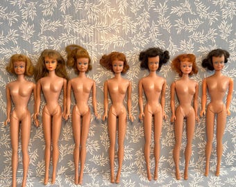 May include: Seven vintage dolls with various hairstyles and skin tones are arranged in a row. The dolls have blonde, brown, and red hair. They are all nude, with articulated limbs and painted facial features, standing against a patterned background.