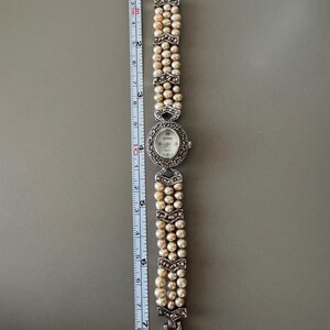 May include: A vintage wristwatch with a pearl bracelet. The watch face is oval with a silver bezel and small hour markers. The bracelet is made of three strands of pearls, accented with silver-toned decorative elements. The watch measures approximately 7 inches in length.