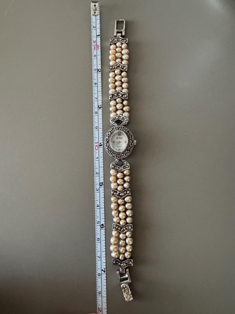May include: A vintage wristwatch with a pearl bracelet. The watch face is oval with a silver bezel and small hour markers. The bracelet is made of three strands of pearls, accented with silver-toned decorative elements. The watch measures approximately 7 inches in length.