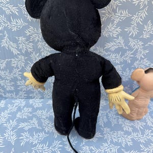 May include: Vintage Mickey Mouse plush toy, viewed from the back. The black plush figure has large ears, yellow gloves, and a long tail. The toy is on a light blue surface with a white leaf pattern.