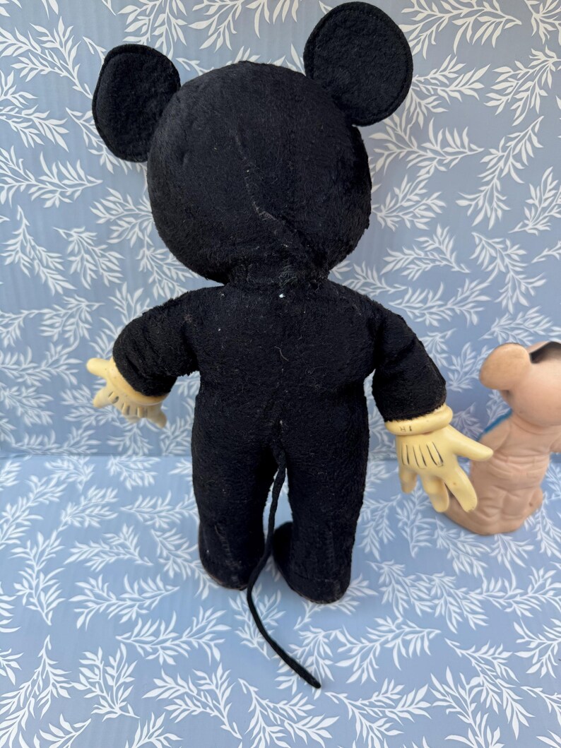 May include: Vintage Mickey Mouse plush toy, viewed from the back. The black plush figure has large ears, yellow gloves, and a long tail. The toy is on a light blue surface with a white leaf pattern.
