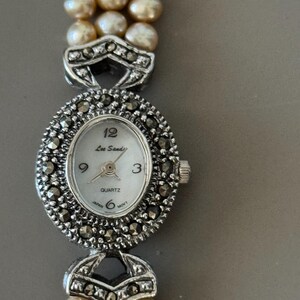 May include: A vintage Lee Sands quartz watch with a silver-tone oval face and a pearl bracelet. The watch face has black Roman numerals and hands. The watch band is made of three strands of pearls. The watch is marked "Japan" and "6 MOVT".