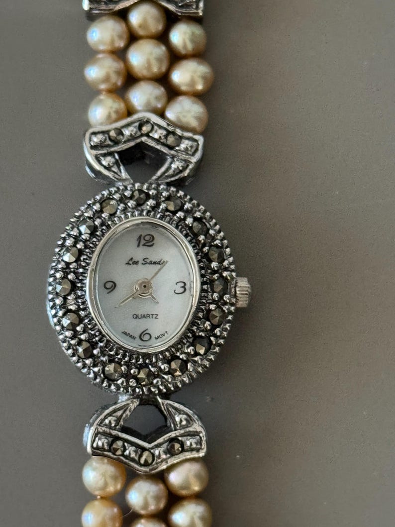 May include: A vintage Lee Sands quartz watch with a silver-tone oval face and a pearl bracelet. The watch face has black Roman numerals and hands. The watch band is made of three strands of pearls. The watch is marked "Japan" and "6 MOVT".