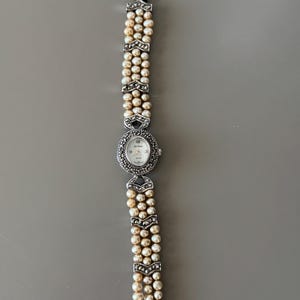 May include: A silver-toned wristwatch with a pearl bracelet. The watch face is oval-shaped, surrounded by small, decorative stones. The bracelet features three strands of pearls, connected by silver-toned, chevron-shaped links.