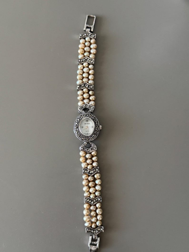 May include: A silver-toned wristwatch with a pearl bracelet. The watch face is oval-shaped, surrounded by small, decorative stones. The bracelet features three strands of pearls, connected by silver-toned, chevron-shaped links.