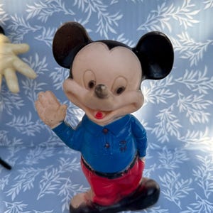 May include: Vintage Mickey Mouse figurine. The figure is wearing a blue shirt, red pants, and black shoes. Mickey's face is light-colored with black ears and a red mouth. The figurine is waving with one hand.