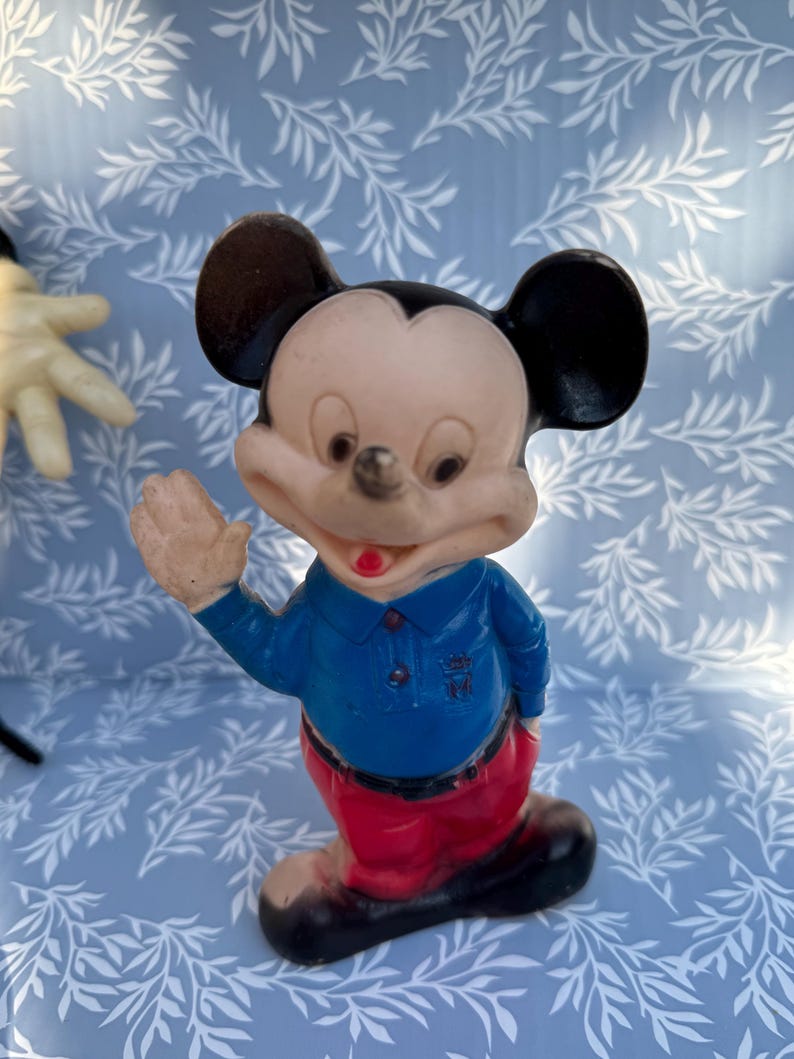 May include: Vintage Mickey Mouse figurine. The figure is wearing a blue shirt, red pants, and black shoes. Mickey's face is light-colored with black ears and a red mouth. The figurine is waving with one hand.